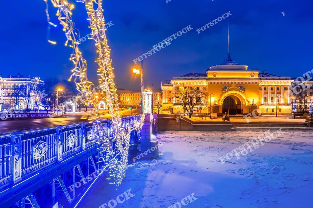 На фотографии изображено здание Адмиралтейства с его знаменитым шпилем, ярко подсвеченное в ночное время. На переднем плане Дворцовый мост, украшенный гирляндами, которые придают праздничную атмосферу. Замерзшая Нева с снежным покровом добавляет ощущение зимней сказки. Этот вид демонстрирует историческую красоту Санкт-Петербурга в праздничные зимние дни.